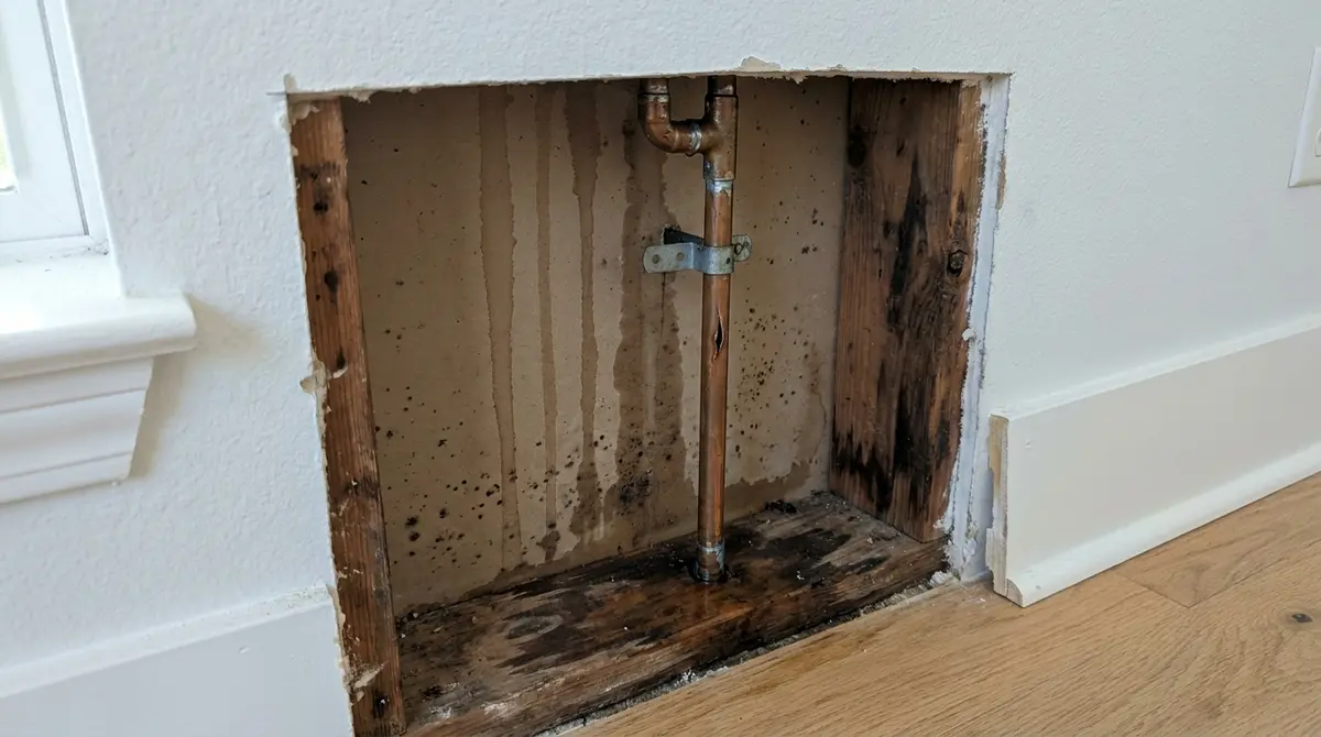 Drywall cut open at the bottom of a wall, showing a copper pipe with a split, water stains and dark mold spots inside the wall cavity