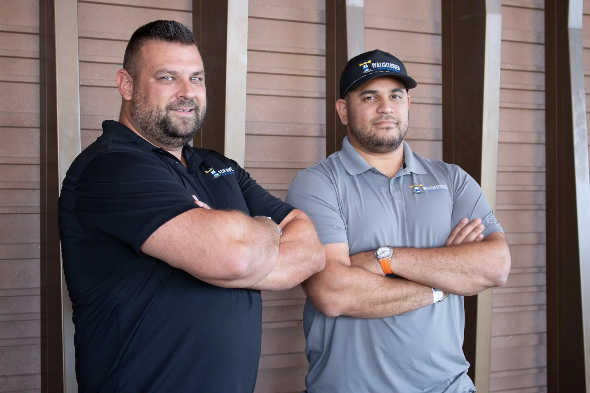 Two team members in black and grey polos posing outside a brown slatted wall, water and fire damage restoration